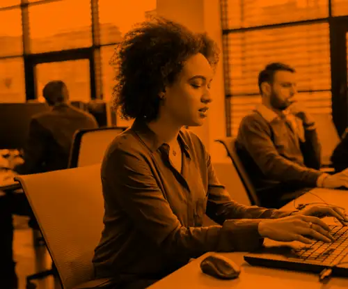 A woman with curly hair sits working on a laptop in an office with several people behind her working in the background.