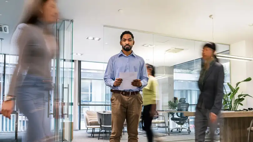 A man stand still in the middle of an office while his co-workers move around him