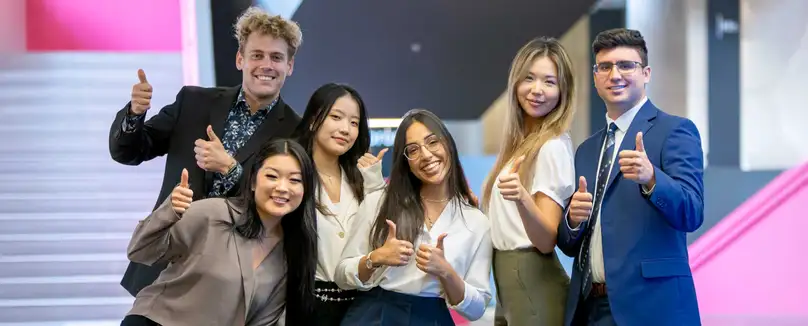 A group of students pose together all giving a thumbs up and standing in front of a pink staircase