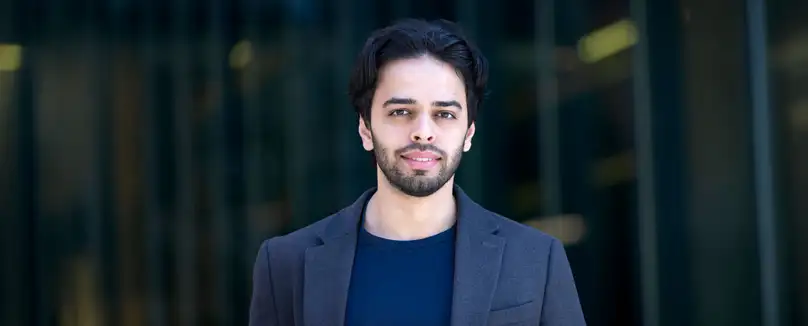 A student with light facial hair stands in front of a black background wearing a grey blazer and a blue crewneck shirt