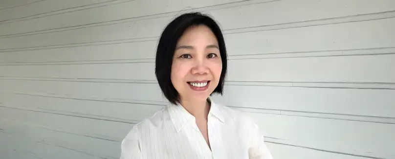 A professional portrait of a dark haired woman in a white blouse standing in front of a panelled white wall