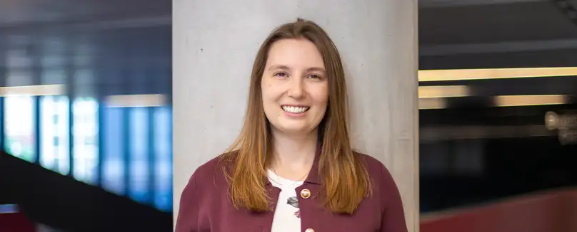A smiling woman in a purple bomber jacket leans against a cement pillar