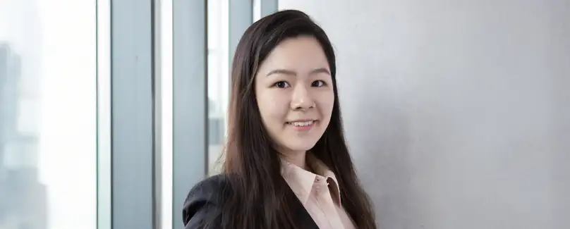 A smiling woman stands in front of a bank of windows. She wears a pink shirt and black blazer.